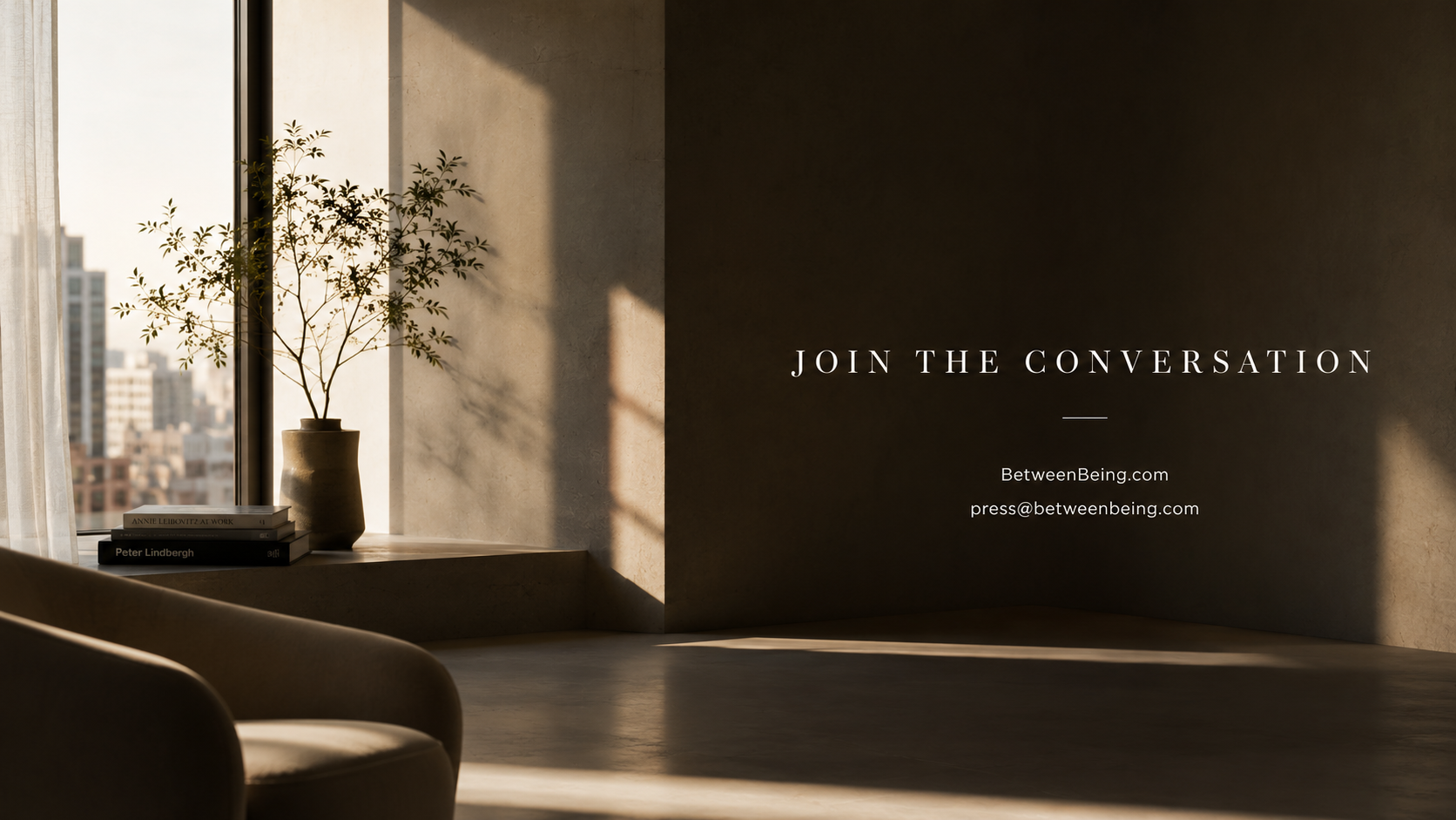 A sunlit minimalist room with a curved armchair, photography books, and a potted tree beside a city window. Text on the wall reads: Join the Conversation — BetweenBeing.com — press@betweenbeing.com